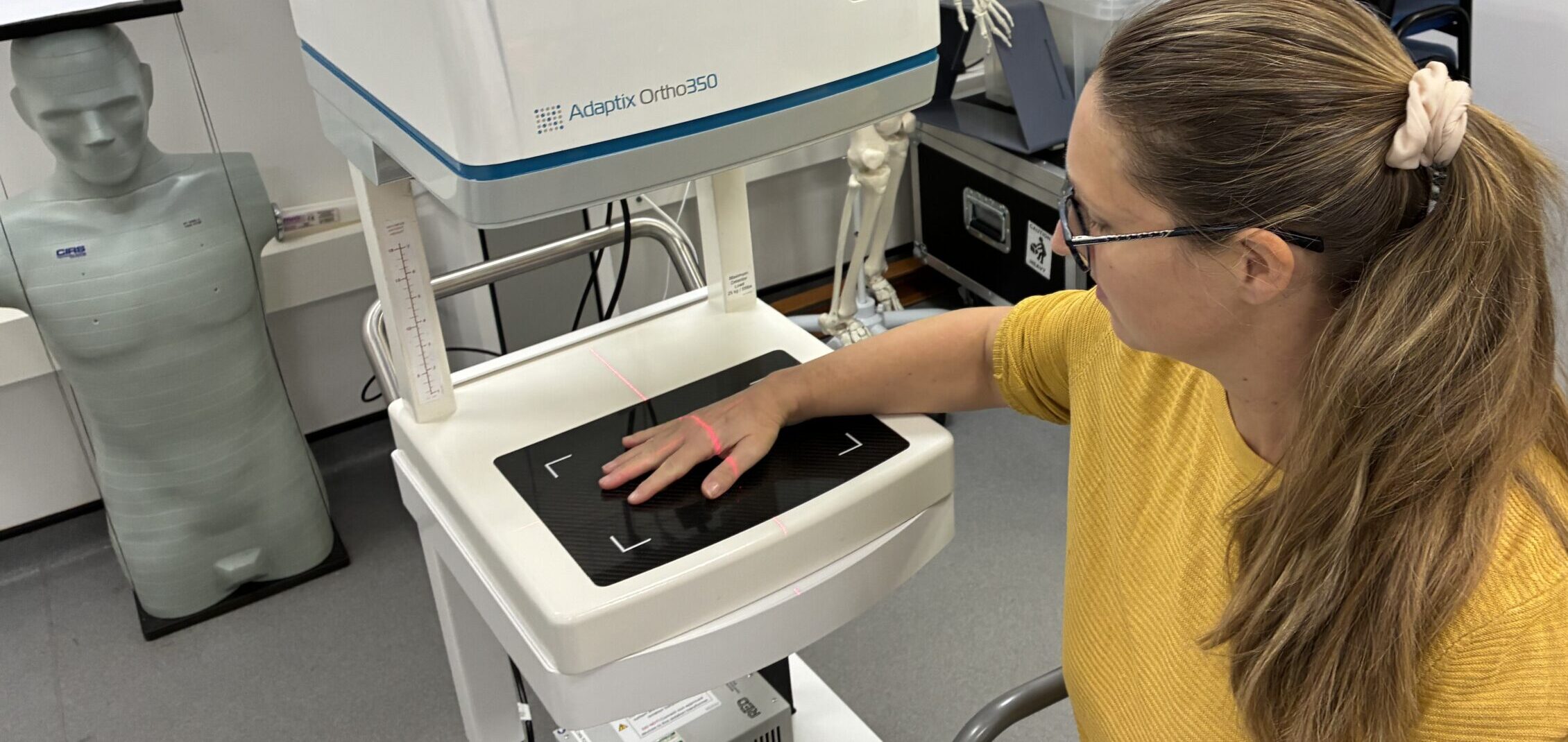 woman sat on chair next to medical imaging machine having hand x-rayed